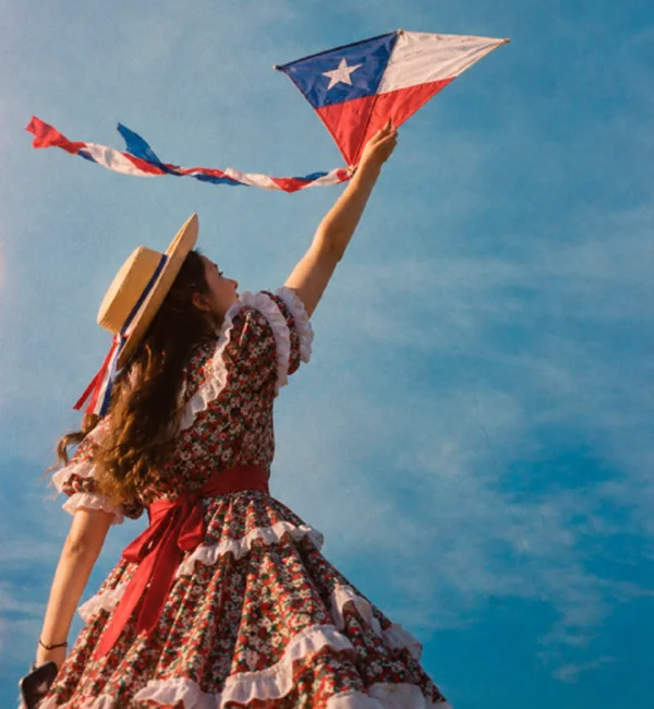 Mujer con vestido de huasa y chupalla elevando un volantín con la bandera de Chile contra el cielo